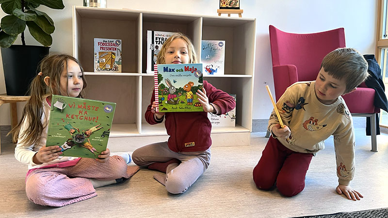 Tre barn sitter på golvet och håller upp varsin bok.